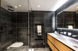 Modern bathroom with dark tile, glass shower, and a double vanity with light wood cabinets.