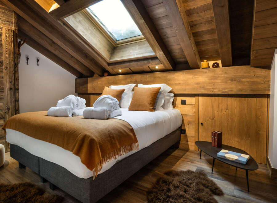 Cozy chalet bedroom with wooden beams, skylights, and a plush bed dressed in warm-toned textiles.