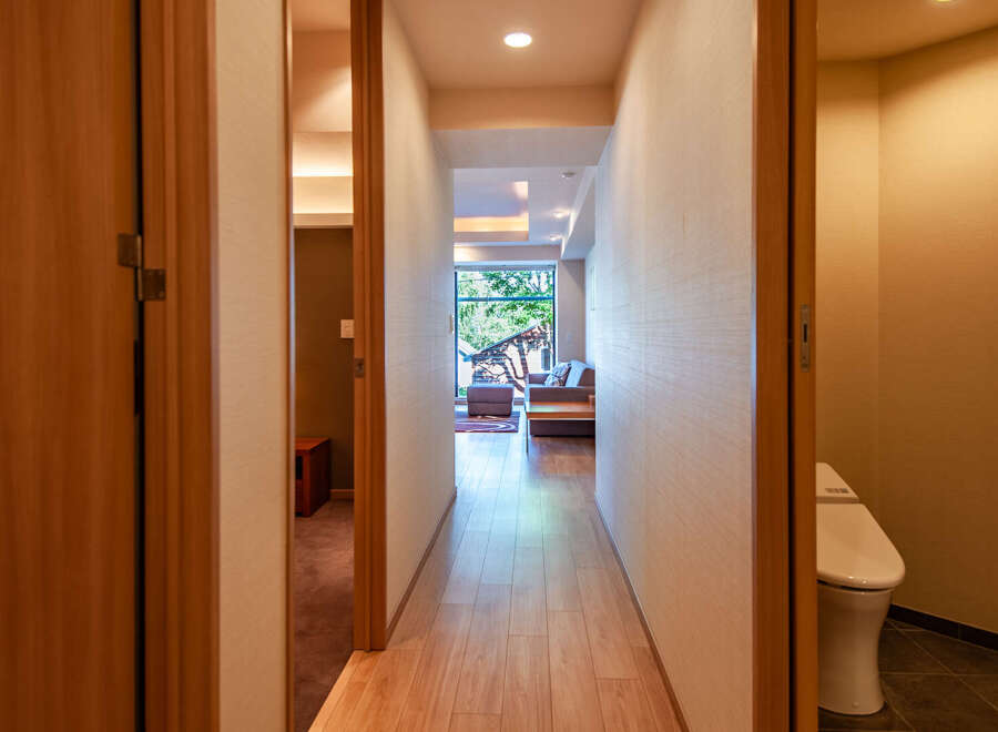 This modern apartment boasts a long hallway with light wood flooring, leading to a living area with large windows and a comfortable sofa.
