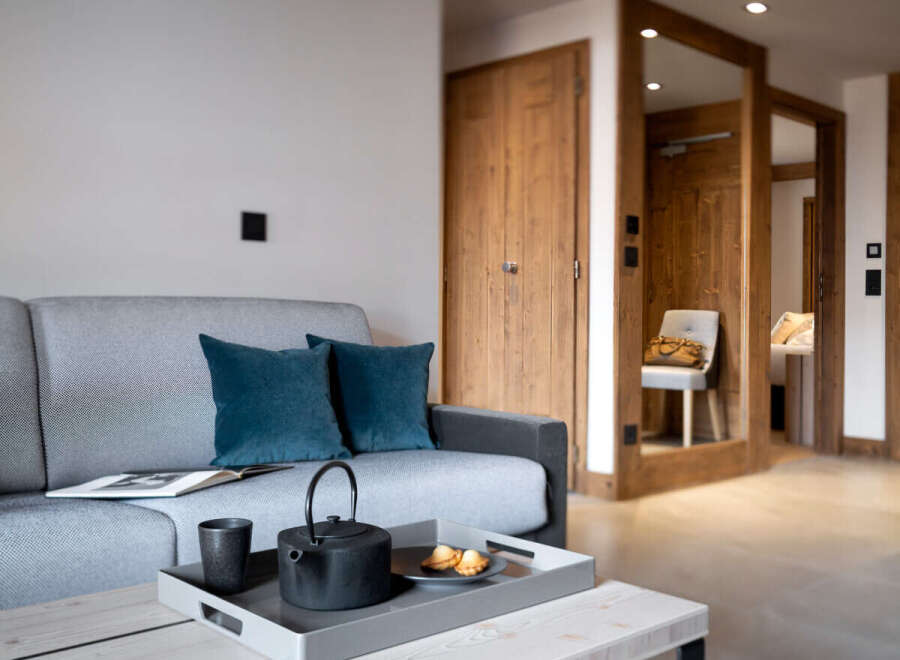 Cozy living room with a grey sofa, teal pillows, and a wooden coffee table featuring a tray with tea set and pastries.