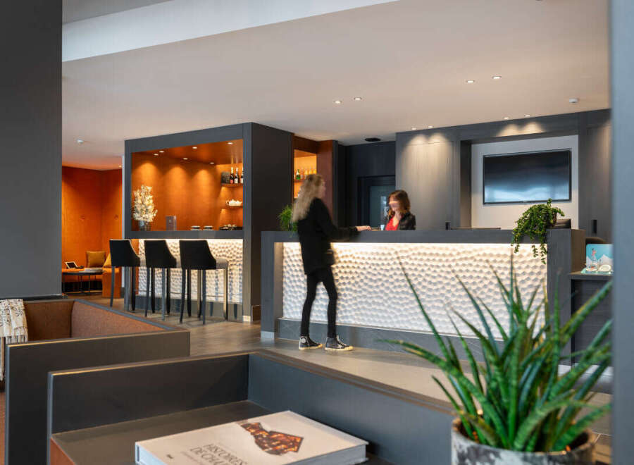 Modern hotel lobby with a stylish reception desk featuring a textured white panel and a built-in bar area with bar stools.