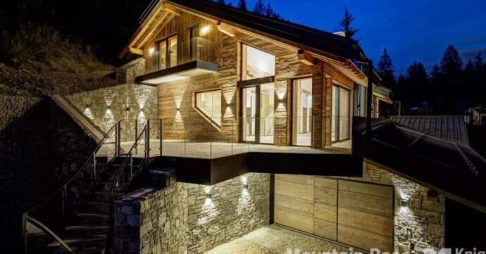 A modern, illuminated wooden chalet with stone accents, set against a nighttime forest backdrop.