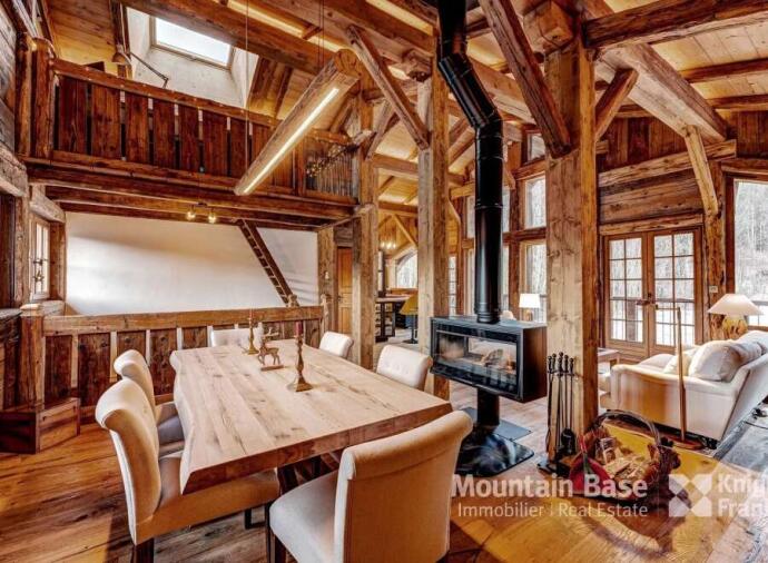 A cozy and rustic chalet interior showcasing a spacious dining area with a large wooden table and upholstered chairs. The room features exposed wooden beams, high ceilings, and a lofted second level. A modern freestanding fireplace with a black chimney serves as a focal point, with natural light streaming through large windows and glass doors. Comfortable seating and warm wood tones create an inviting ambiance.