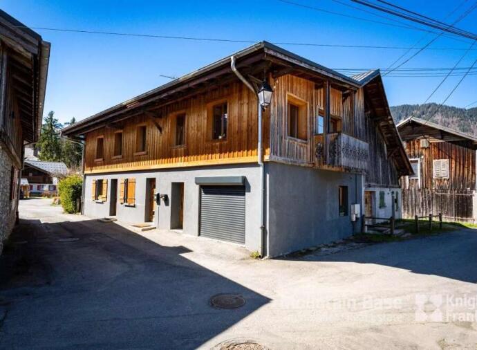 Two-story rustic house with wooden upper facade and gray lower walls, situated on a quiet village street.