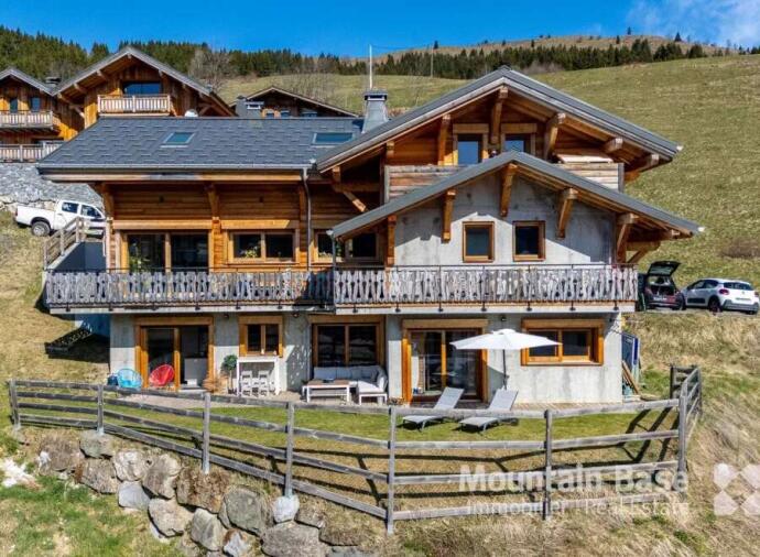 Large chalet-style house with wooden and concrete exterior, multiple balconies, and outdoor seating, set on a grassy hillside.