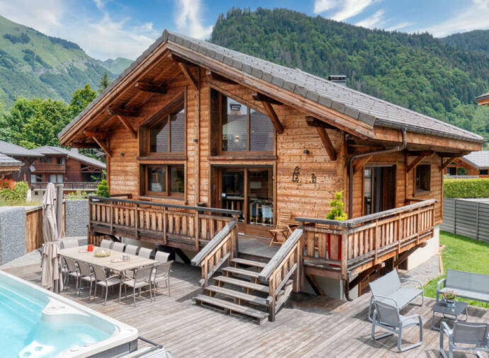 Cozy wooden chalet with outdoor seating and hot tub, set in a green mountain landscape.