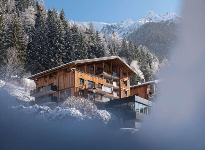 A wooden chalet with balconies in a snowy mountain area, surrounded by pine trees and snow-covered peaks.