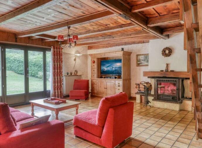 Cozy living room with wooden beams, red furniture, fireplace, TV, and large windows with red curtains overlooking a green lawn.