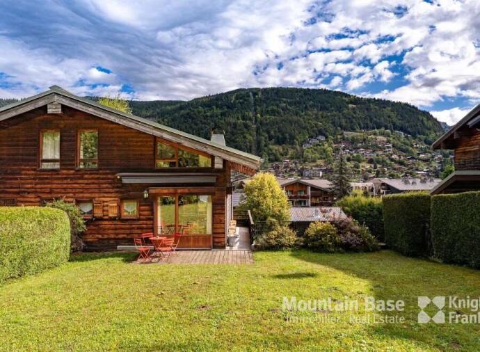 A wooden chalet with large windows and a patio in a green yard, surrounded by trees and mountains; logos of Mountain Base Immobilier Real Estate and Knight Frank are in the corner.
