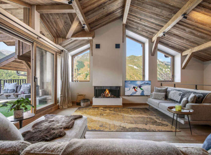 Modern chalet living room with wooden beams, sofas, fireplace, and mountain views through large windows.