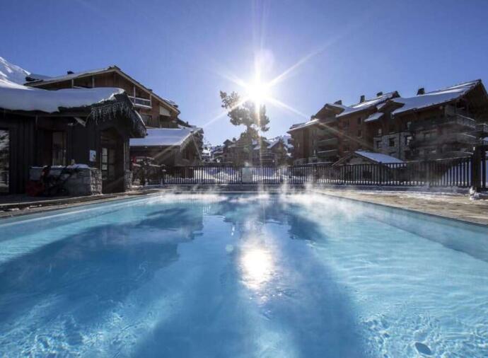 Outdoor heated pool with steam rising, surrounded by snow-covered mountain chalets under bright sunlight and clear blue sky.