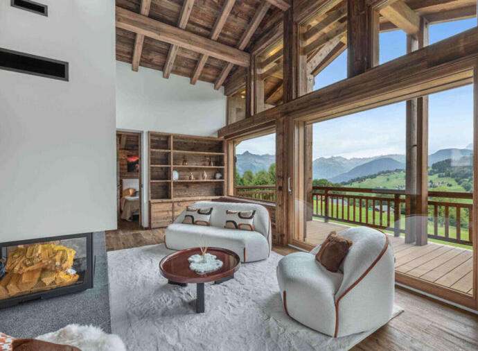 Modern mountain chalet living room with wooden beams, large glass windows, cozy white armchairs, and scenic view of green hills and mountains.
