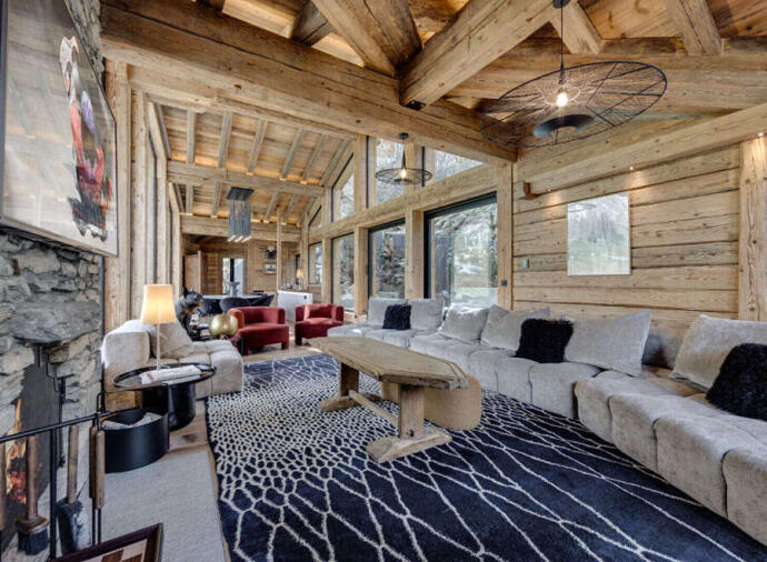 Rustic mountain chalet living room with exposed wooden beams, large windows, cozy sectional sofa, modern lighting, and patterned rug.