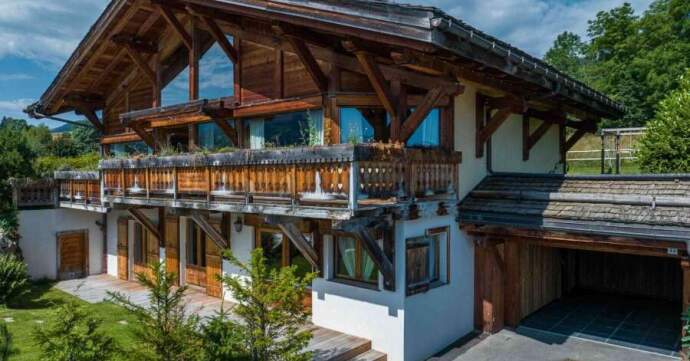 A beautiful alpine chalet-style house featuring a wooden facade with exposed beams, large windows, and a wraparound balcony. The property includes a garage, a paved driveway, and a well-maintained lawn with lush greenery, set against a backdrop of trees and a partly cloudy sky
