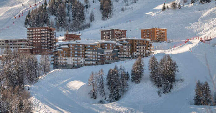 Snow-covered ski resort with modern chalet-style buildings nestled on a mountainside, surrounded by ski slopes, evergreen trees, and skiers on the trails.