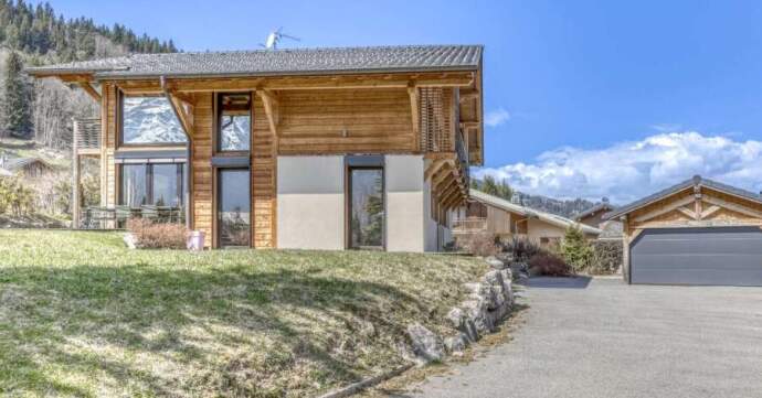 Modern wooden chalet with large windows and sloped roof, set on a grassy hill with mountains in the background.