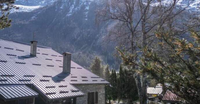 A stone house with a metal roof and two chimneys in a mountainous area, surrounded by trees with snow-capped peaks in the background.
