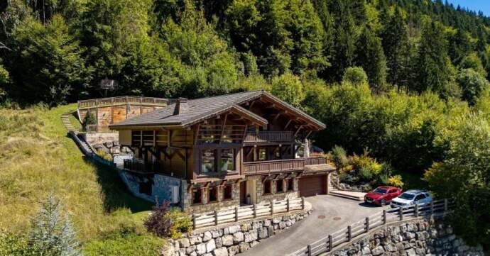 A large wooden chalet-style house with balconies and a stone foundation, nestled on a green hillside with two cars parked in the driveway; logos of Mountain Base and Knight Frank appear in the corner.