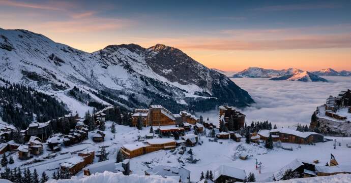 Snow-covered Alpine ski resort village at sunset, with wooden chalets overlooking mountain peaks and a cloud inversion in the valley below.